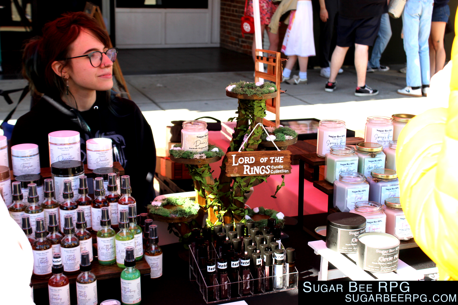 A woman with reddish brown hair and glasses and white skin sits at a table selling scents, bath scrubs, candles, and lotions.  There is a display for Lord of the Rings candle sets in the middle. 

Sugar Bee RPG
SugarBeeRPG.com