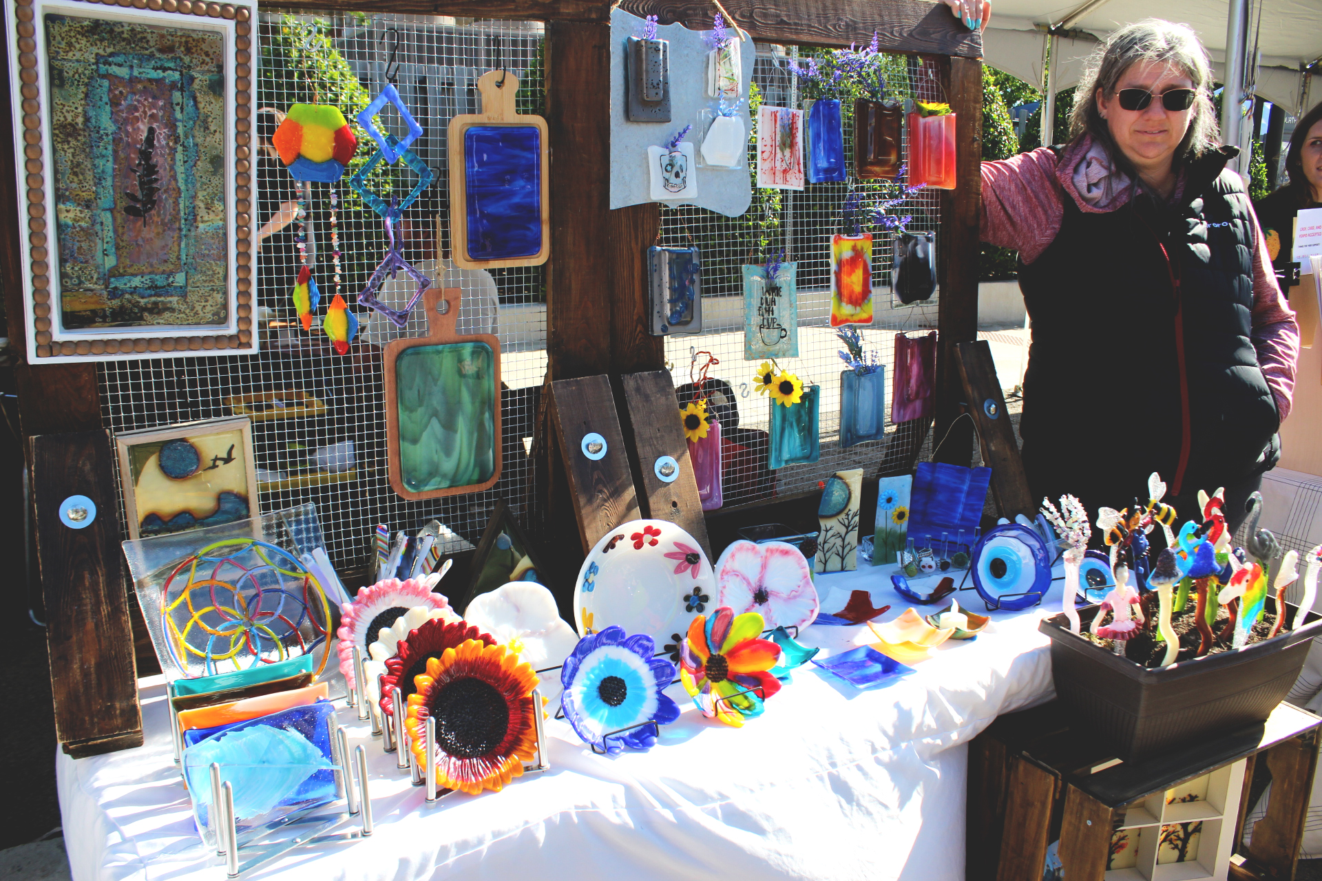 An older woman with white skin and grey hair is seen wearing sunglasses displaying her glassware work that ranges from plates to hanging decor to garden decor. 