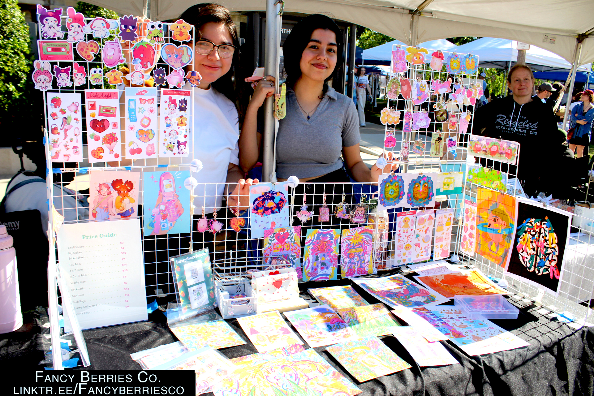 Two people standing at their booth featuring cute anime and cartoon style prints and stickers. 