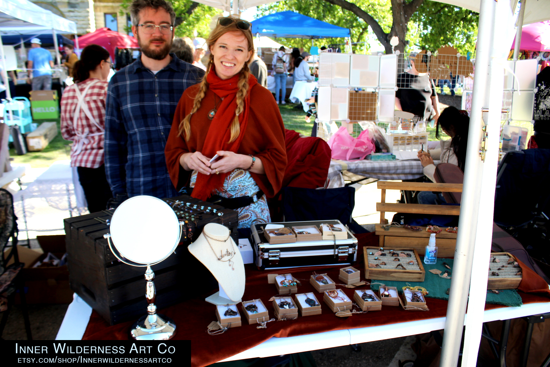 A married couple standing behind their booth with a man in a plaid blue shirt with white skin and glasses and a woman in a red outfit with scarf and long blond braided hair and white skin. The wares include wire copper jewelry and decorative items. 