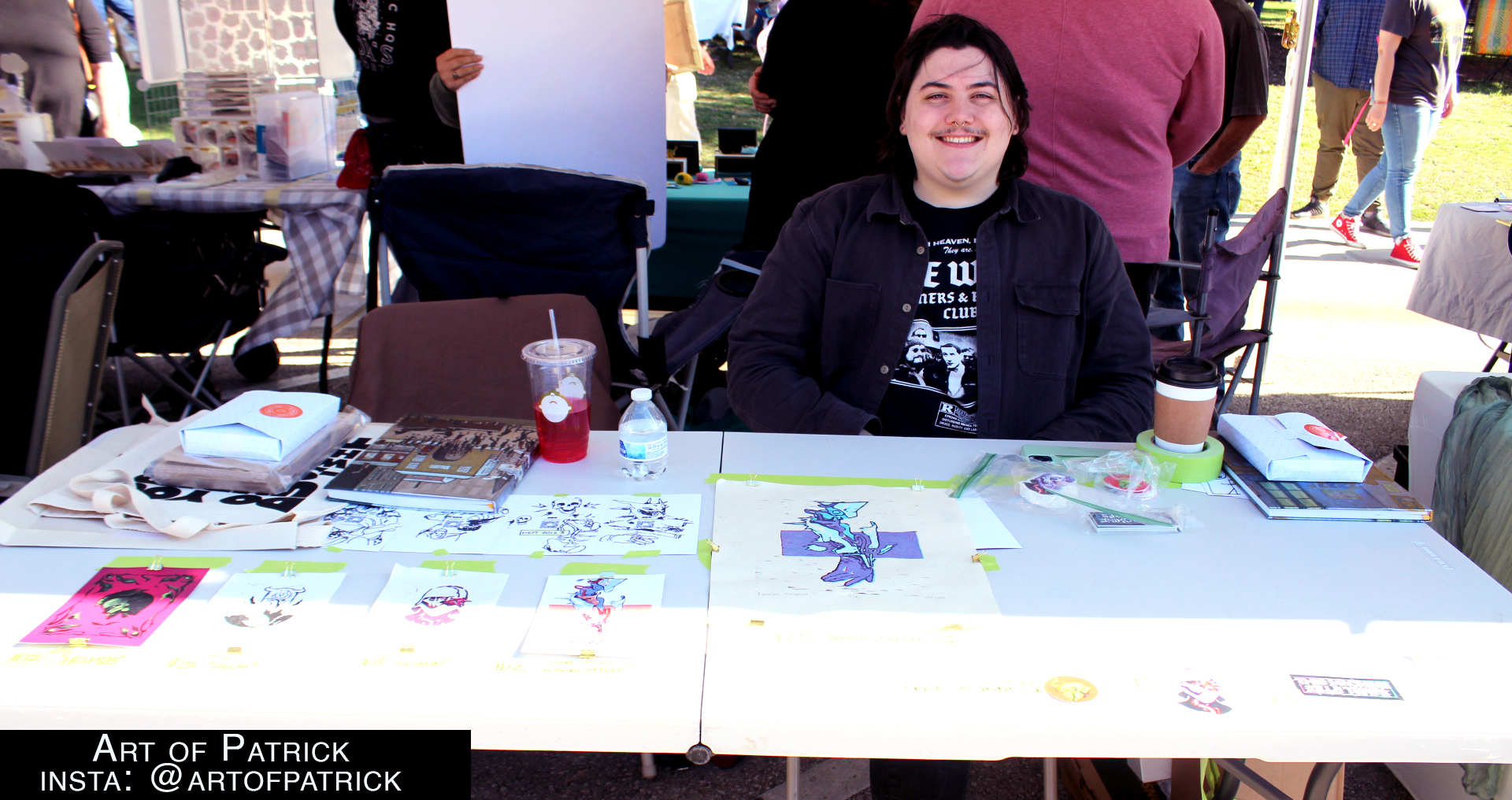 A white skinned nonbinary man with black hair is wearing all black and is sitting at their table displaying their art.