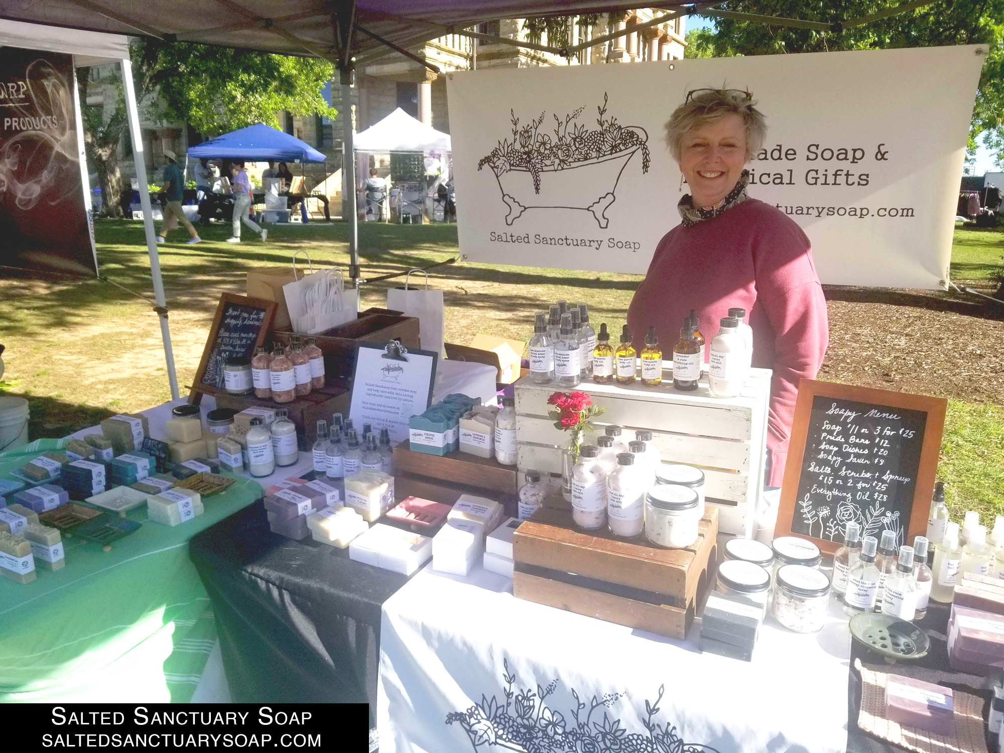 A white woman with short grey blonde hair is wearing a crimson sweatshirt and has soaps and bath goods for sale on diisplay. 
