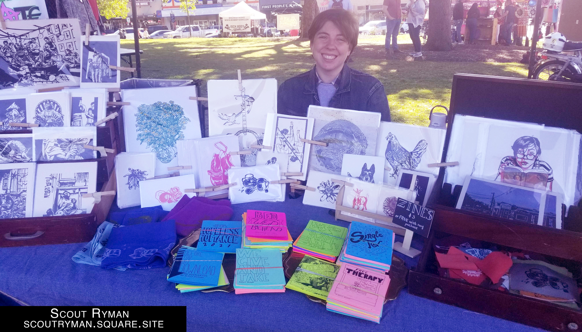 A person with white skin and short brown hair is displaying their artwork and zines on a blue tabletop. 