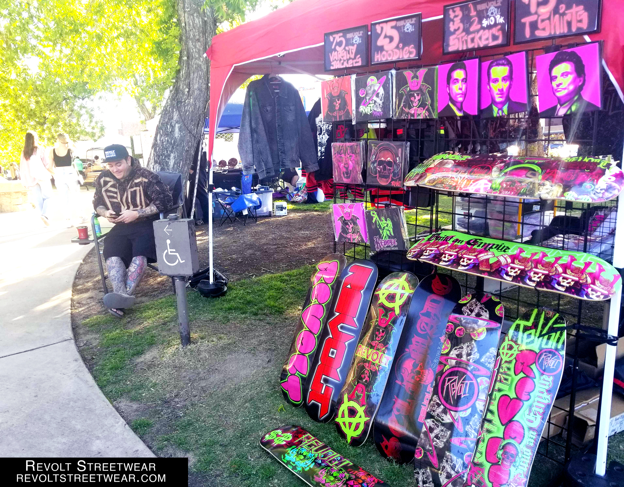 A person is sitting outside their tent with their wares, Revolt Streetwear uses lime and pink heavily in their skater inspired clothing and boards. 