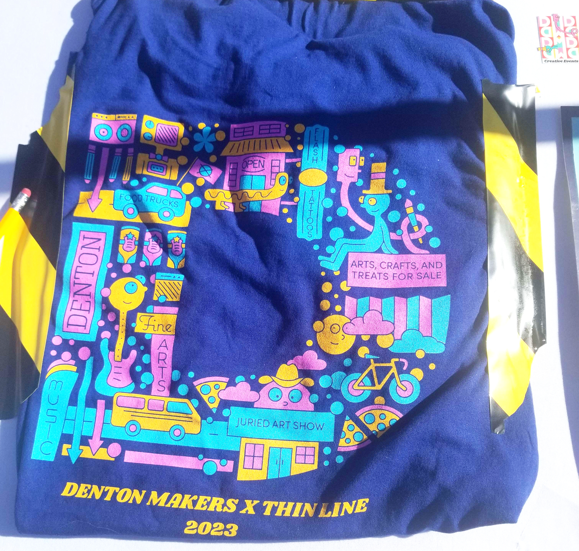 Denton Maker's Fest T shirt design is a large D made out of multiple cute style buildings notably in Denton, trucks, people, businesses, tattoo parlors, guitars, instruments bikes, pizza, etc. 

Says at bottom:

DENTON MAKERS X THIN LINE [FEST for both]
2023
